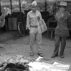 Merchant trying to make a sale of cloth to a soldier who has a plastic carrying sack