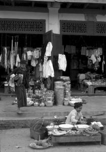 Shop featuring pails, kettles, umbrellas, shirts; Lao woman with food stall on street in front of shops