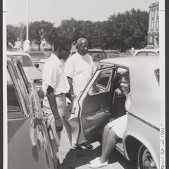 Two men stand next to a car where a woman sits