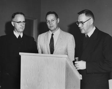 Three unidentified men standing by lectern.