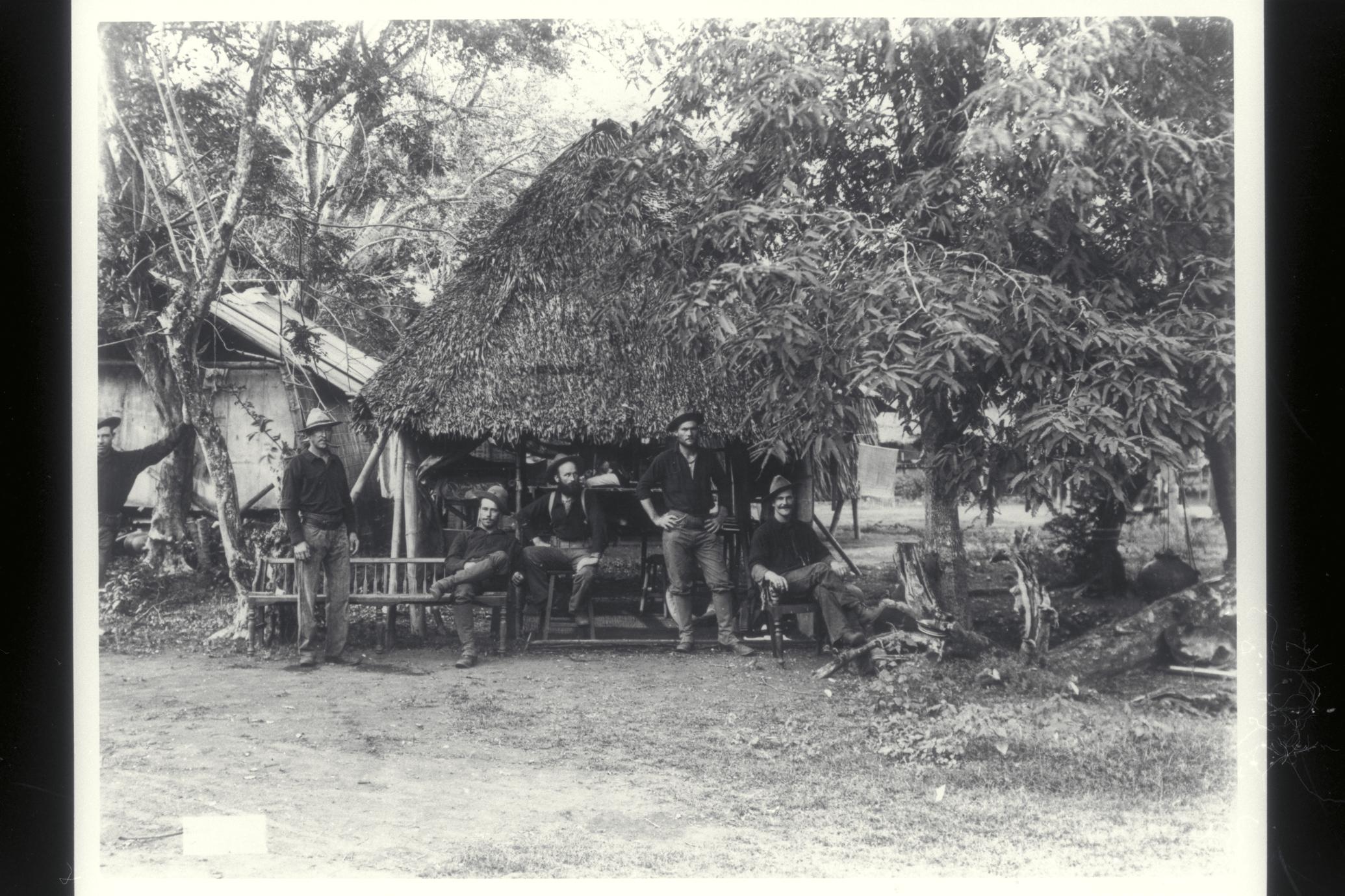 Six U.S. soldiers relax next to a thatched hut and shade trees, 1899