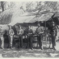 Six U.S. soldiers, arranged in a line according to their height, pose with their rifles next to a thatched and tin-roofed building, 1899