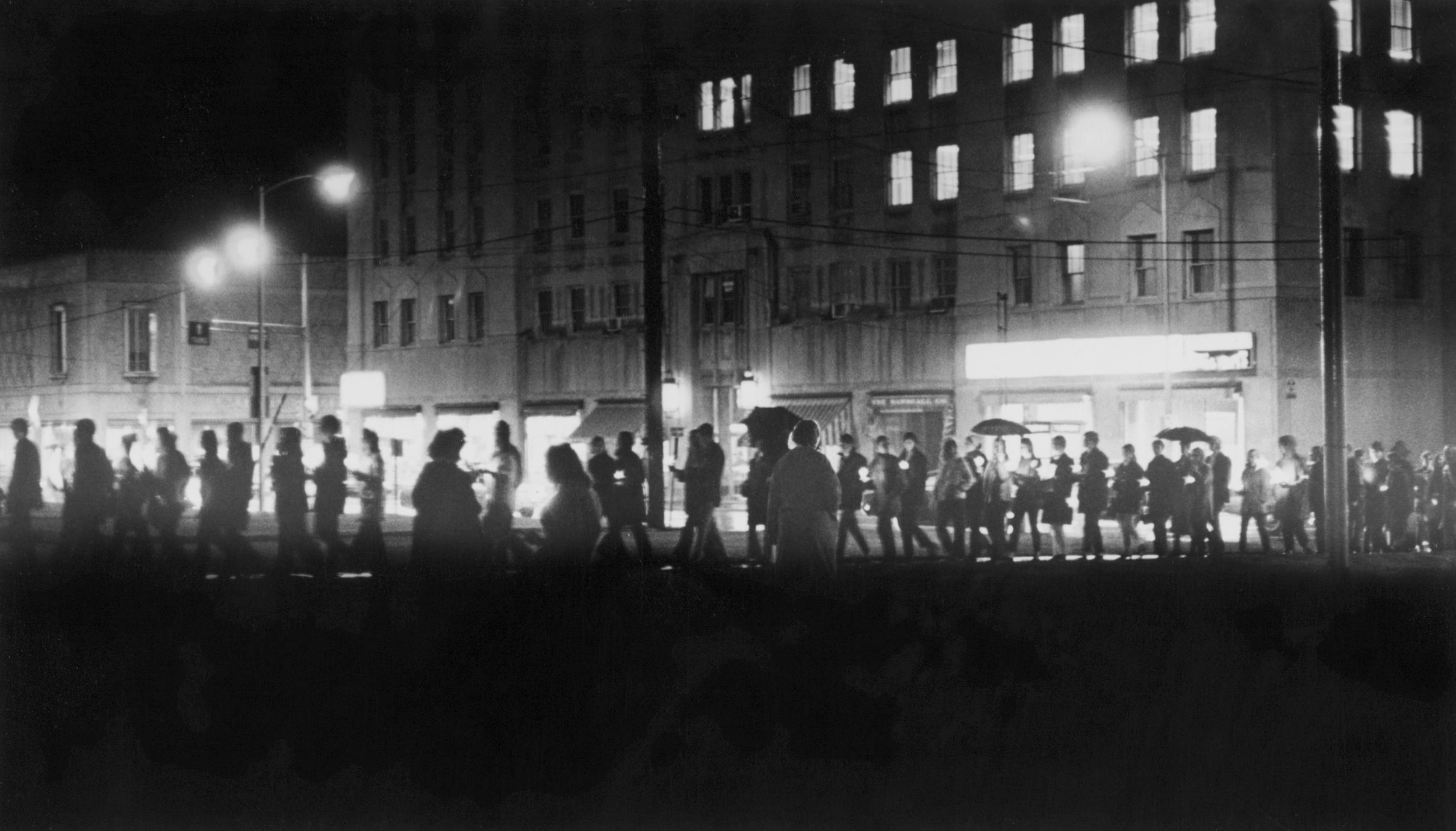 A black and white photo of people holding umbrellas marching through the darkness by candlelight