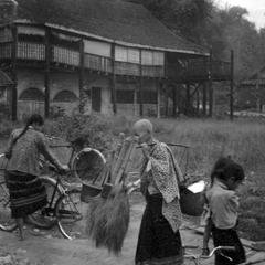 Lao woman with shaved head carrying brooms and rice to market, a younger Lao woman gets on her bicycle
