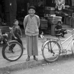 Old Chinese merchant in front of clothing shop, children in background