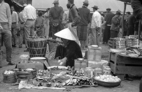 Vietnamese woman merchant selling fish on banana leaves, eggs, onions, with baskets of charcoal at her back, groups of men in background looking at merchandise