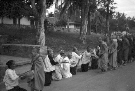 Old women with shaved heads on bended knees offering food to monks