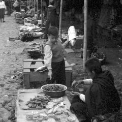 Line of women vendors with stands, foreground woman selling rice cakes and fried bananas