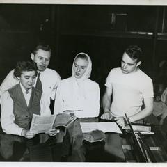 Four Stout Symphonic Singer members standing around a piano singing from a song book