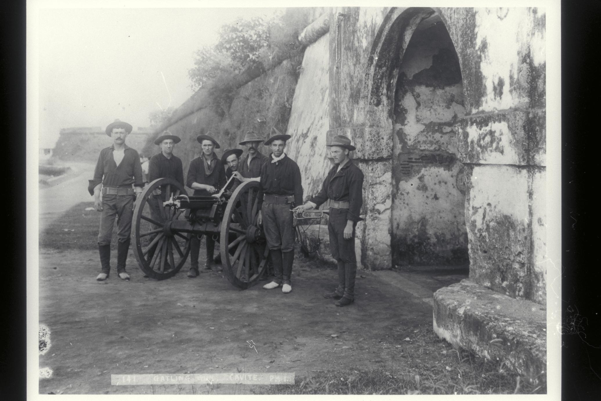 U.S. soldiers pose next to a Gatling gun next to the city walls, Cavite, 1899