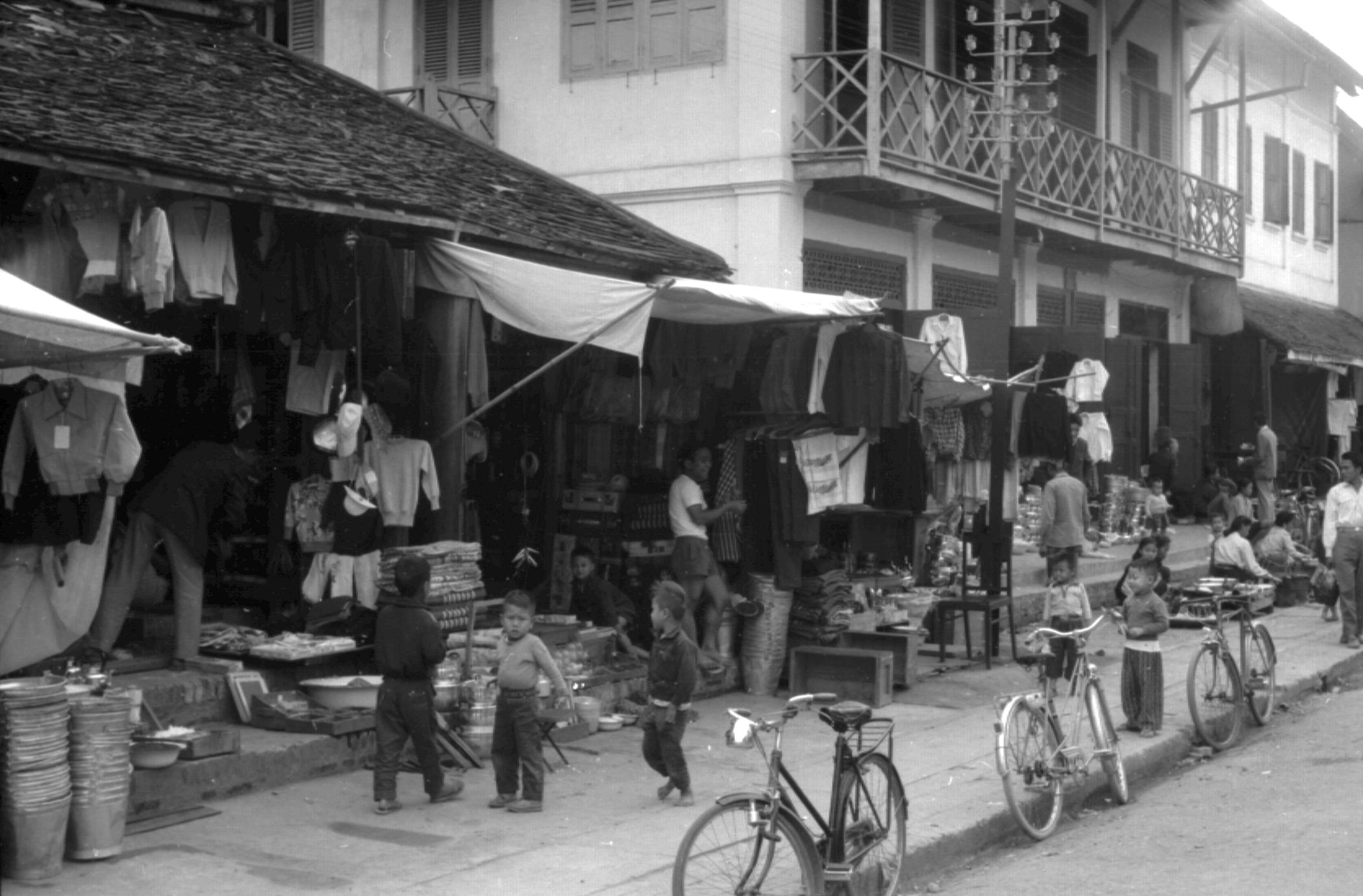 View of a number of shops along main street, bicycles lined up, selling tin pails, clothing, electric pole visible, upper story probably used for residence