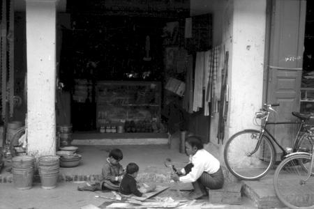 Young woman with merchant's children playing outside shop; galvanized tin pails on left