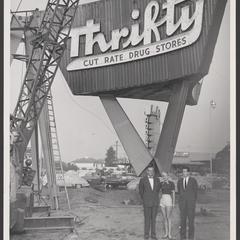 Three people stand outside at a construction site next to a large drugstore sign