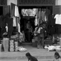 Chinese shop with household utensils, clothing, umbrellas; women in front prepare cooked food for sale