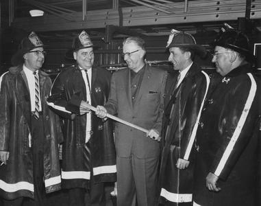 Four men in firemen's gear and one man holding a fire axe are standing in the fire station.