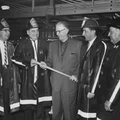Four men in firemen's gear and one man holding a fire axe are standing in the fire station.
