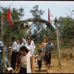 Inauguration of United States Operations Mission (USOM) dam at end of procession, women with children passing under palm framed archway with Lao and United States flags