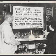 A pharmacist and a shopper view a warning sign for prescriptions