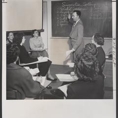 Man stands at a chalkboard talking to a group of seated women