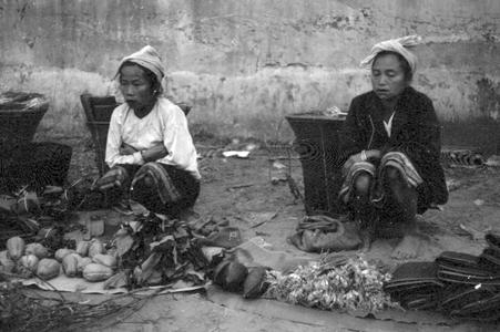 Kammu (Khmu') women crouching near their forest products displayed for sale with baskets in background against wall