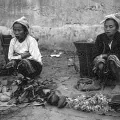 Kammu (Khmu') women crouching near their forest products displayed for sale with baskets in background against wall