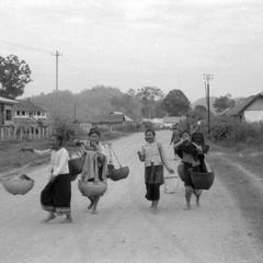 Barefoot Lao villagers returning home after market with baskets on shoulder poles, homes on stilts on left, poles carrying electricity and perhaps phone lines