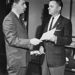 Two men standing in an office exchanging a piece of paper.