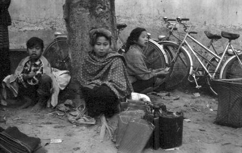Kammu (Khmu') woman at market with produce wrapped in banana leaves leaning against tree, sitting on ground, local woman in back with her bicycle
