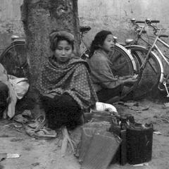 Kammu (Khmu') woman at market with produce wrapped in banana leaves leaning against tree, sitting on ground, local woman in back with her bicycle