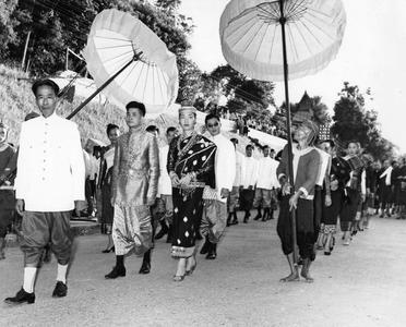 After ceremony, newlyweds walk through main street of town, shaded by white silk royal umbrellas held by Kammu (Khmu') honor guards